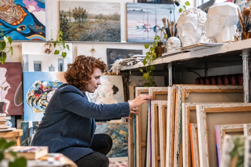 Artist organizing unsold paintings and canvases in a working studio.