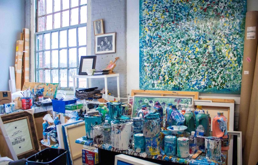Cluttered artist studio filled with paint-splattered cans, unfinished canvases, and large abstract paintings.