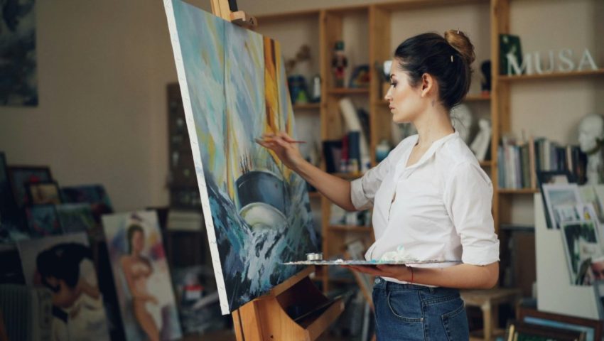 Artist painting in a studio surrounded by canvases.