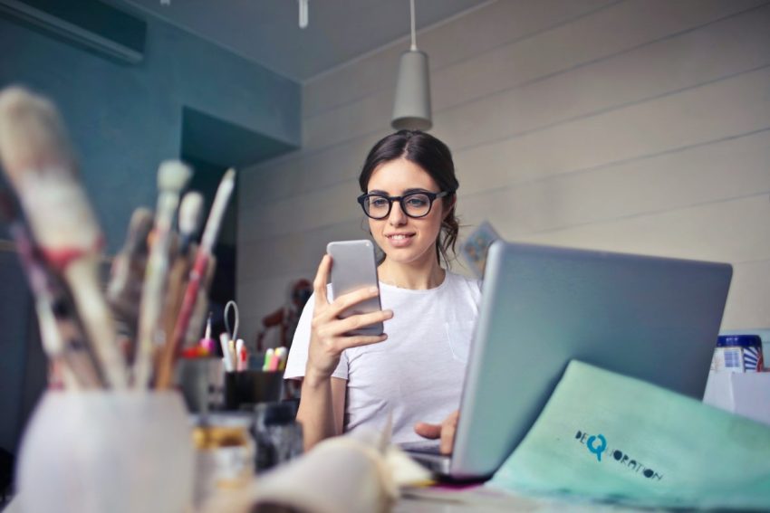 Artist managing creative business work on a laptop and smartphone surrounded by paintbrushes.