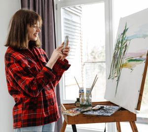 An artist holding a smartphone to photograph her painting on an easel