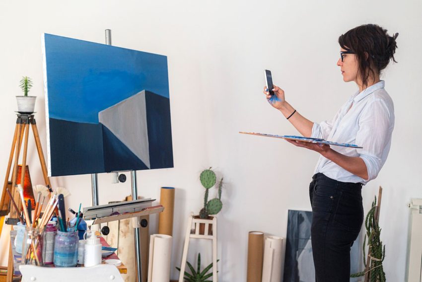 An artist in a studio taking a photograph of her geometric abstract painting with her phone while holding a palette.