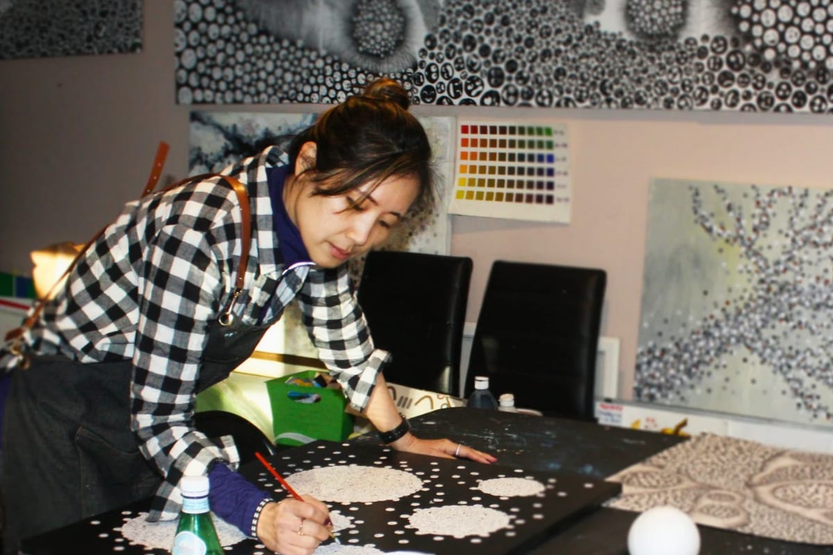Artist Sarah Shinhyo Kim painting a detailed dot-pattern canvas in her studio, with her finished works covering the walls