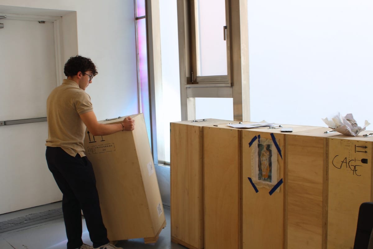 Agora Gallery staff member moving a wooden shipping crate marked with orientation arrows beside a row of labeled art crates