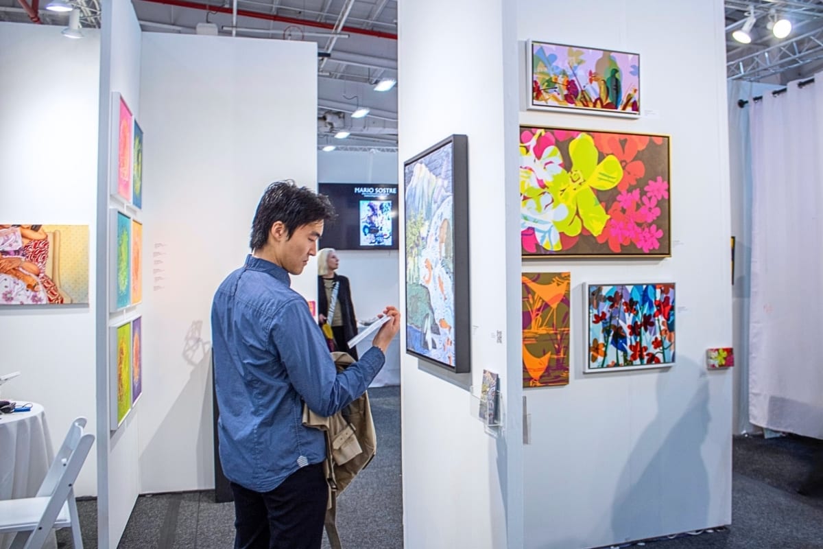 A visitor reads an artist card beside colorful floral paintings at the AGI Fine Art booth at Artexpo New York.